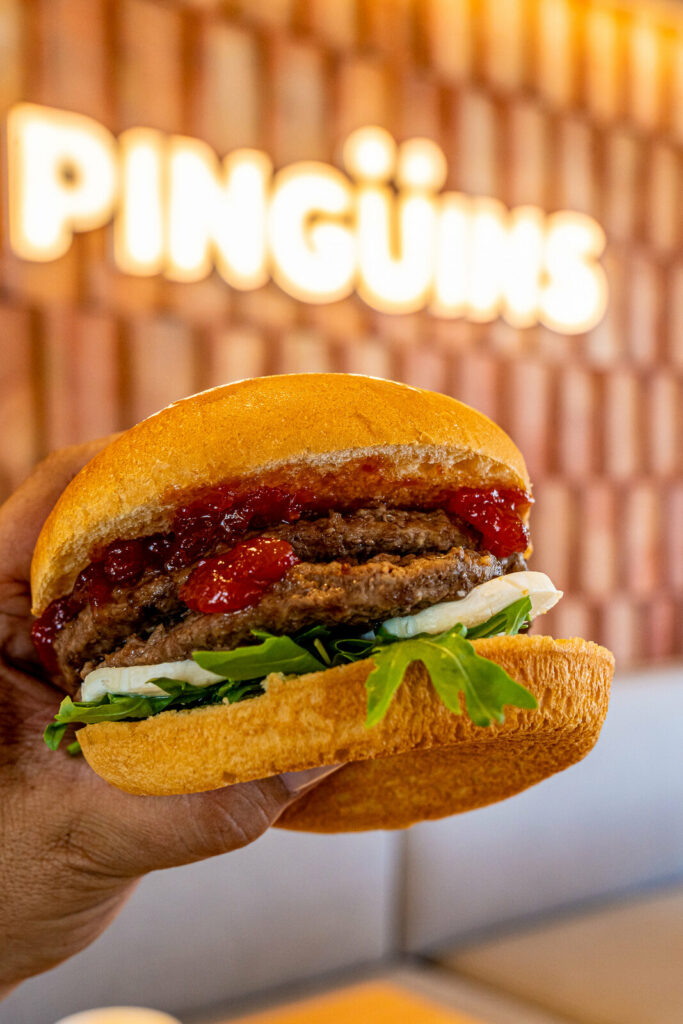 Burger en la mano con el rótulo iluminado de Pingüins al fondo