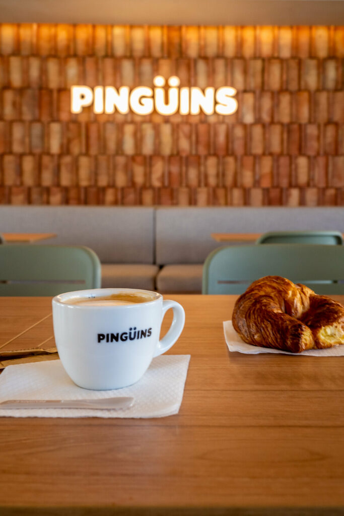 Café en taza de Pingüins con croissant y el rótulo iluminado al fondo